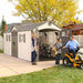 Lifetime 20' Cottage Style Plastic Shed, Dual Entrance, front and right view of building showing the two entrances with double doors and two windows with shutters.  Right side (end) doors are held open by a woman with a man and boy bringing out a ride on mower and leaf blower, showing the scale of size and what you can fit in the building.