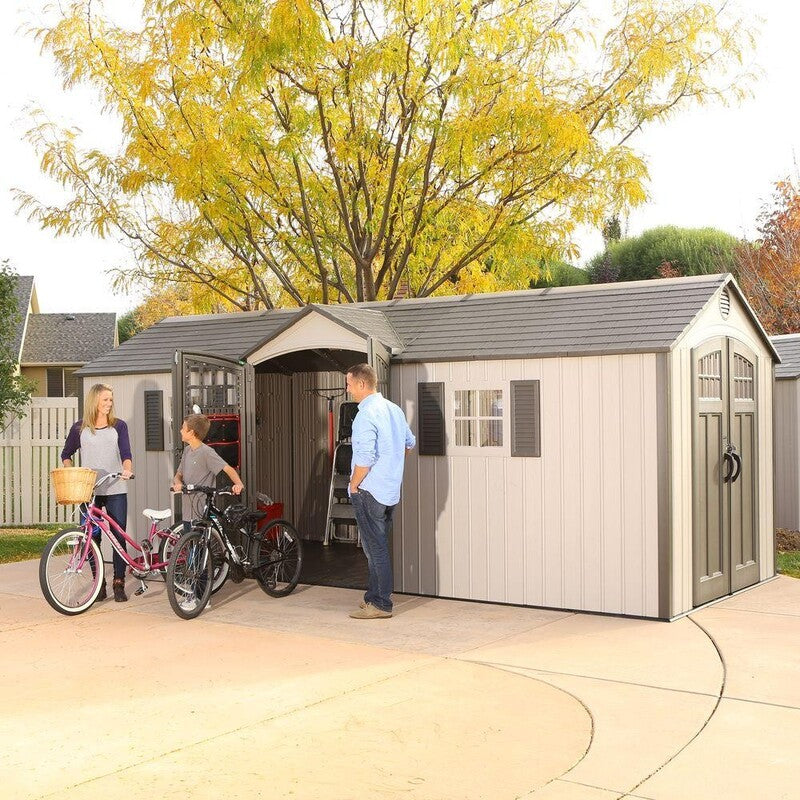 Lifetime 20' Cottage Style Plastic Shed, Dual Entrance, front and right view of building showing the two entrances with double doors and two windows with shutters.  Front side doors are held open by a man with a woman and boy bringing their bikes out of the building, showing the scale of size and what you can fit in the building.